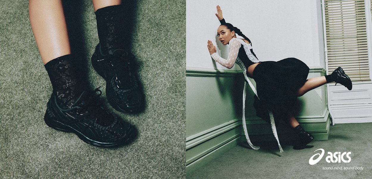 Woman in black lace socks and sneakers poses dynamically against green walled corner, wearing black skirt and white sheer top with braided hair.
