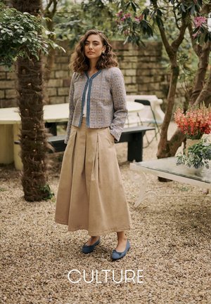 Graublaues Tweed-Jackett mit rundem Ausschnitt, kombiniert mit einem beigen, plissierten Maxirock und blauen Ballerinas, in einer Gartenumgebung im Freien.