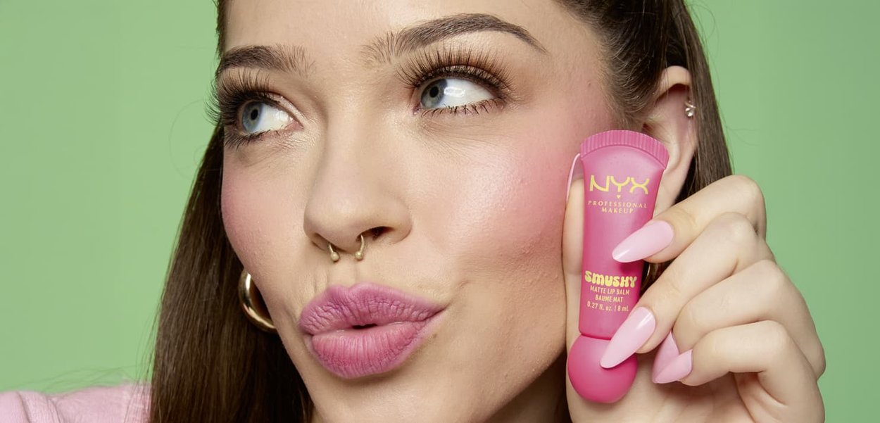 Balsam do ust NYX Smashy Matte w małej różowej tubce z zaokrąglonym końcówką. Modelka ma długie brązowe włosy i różowe paznokcie na tle zielonym.