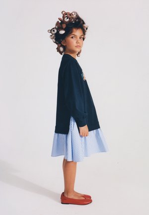 Young girl wearing a navy sweatshirt, blue striped skirt, red flats, and teddy bear hair accessories, standing against a plain white background.