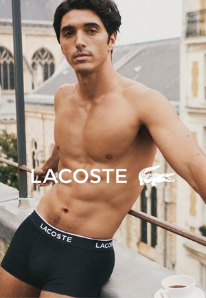 Czarne bokserki Lacoste z białą gumką z logo marki, model z odkrytą górą ciała i ciemnymi włosami średniej długości, pozujący na zewnątrz.