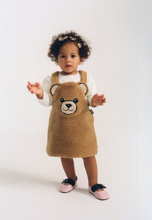 Toddler with curly hair wearing a brown teddy bear dress, white long-sleeve shirt, pink shoes, and bear hair clips standing on a plain background.