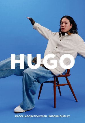 Model trägt eine cremefarbene Jeansjacke über einem schwarzen Shirt und blauen Jeans, sitzt auf einem Holzstuhl vor einem blauen Hintergrund.