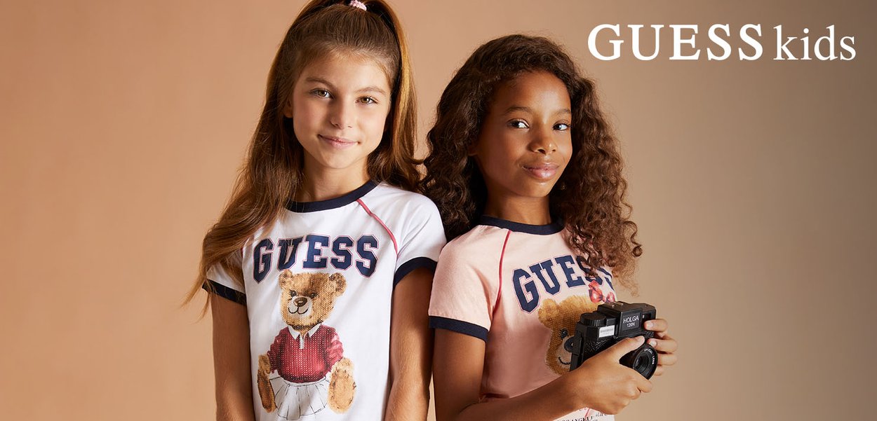 Due ragazze vestite con magliette GUESS Kids con stampe di orsetti, una con una macchina fotografica vintage, posano contro uno sfondo beige neutro.