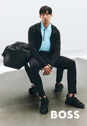 Schwarze Sportjacke über einem hellblauen Poloshirt, schwarze Jogginghosen und Turnschuhe. Große schwarze Sporttasche neben dem sitzenden Modell auf einem grauen Boden.