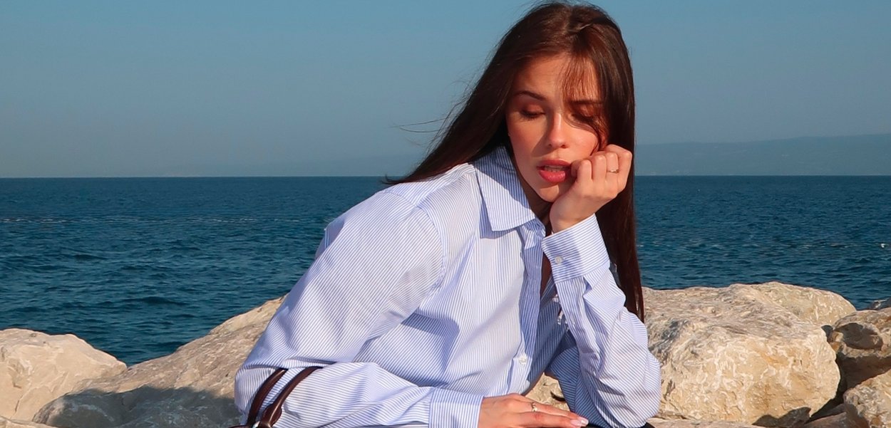 Hellblau-weiß gestreifte Button-Up-Bluse mit langen Ärmeln, lockere Passform, getragen im Sitzen an einer felsigen Küste mit Ozeanhintergrund.