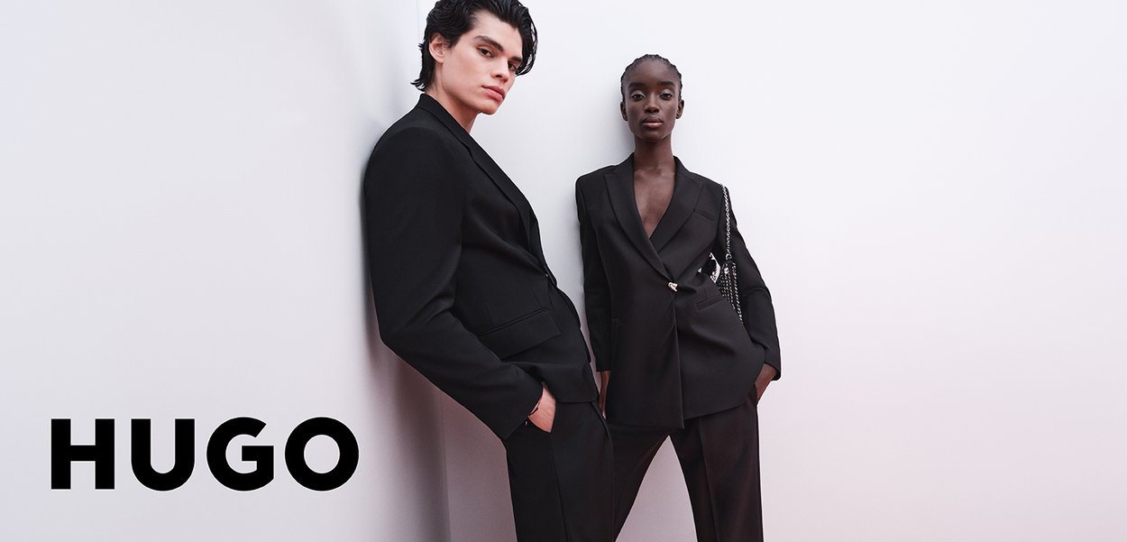 Two models in tailored black suits, standing against a white background. The male suit has a slim fit; the female suit features a single button and accents.