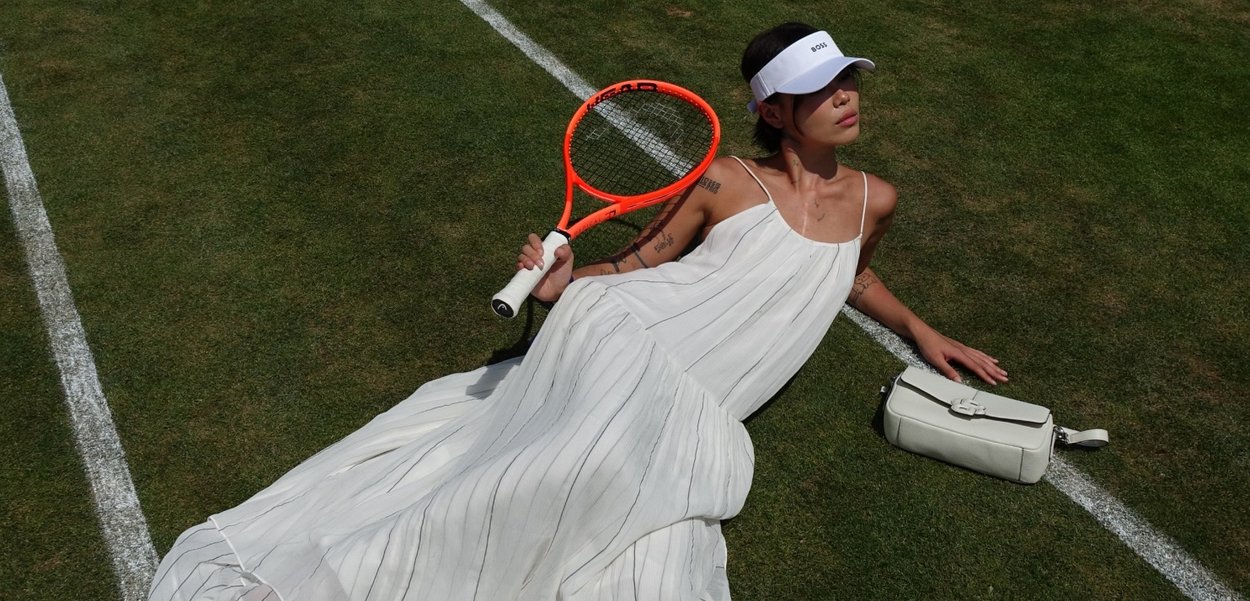 Abito bianco plissettato con spalline sottili, abbinato a una borsa bianca. Racchetta da tennis arancione in mano, su erba verde con linee bianche.