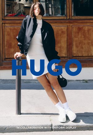 Giubbotto bomber nero, abito di maglia bianco, sneakers bianche e calze bianche. In piedi accanto a un palo contro uno sfondo di legno. Testo blu "HUGO."