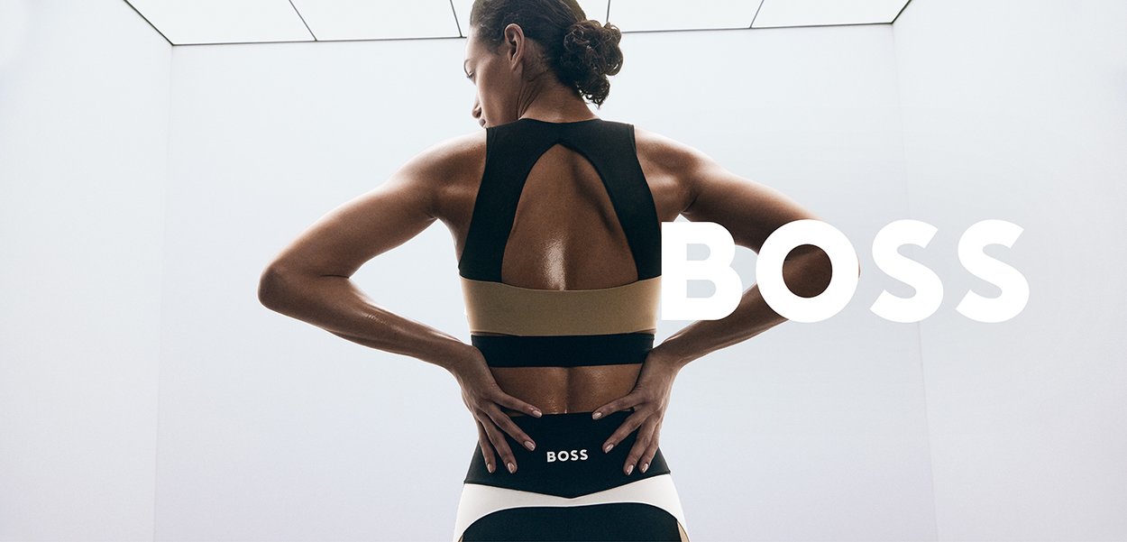 Schwarzer und beiger Sport-BH mit offenem Rückendesign, Logodetail an der Taille, an einem Model, das von hinten gegen einen schlichten Hintergrund blickt.