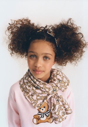 Jeune fille avec des cheveux bouclés volumineux attachés, portant un pull rose ainsi qu'une écharpe et un bandeau à motifs ours en peluche assortis.