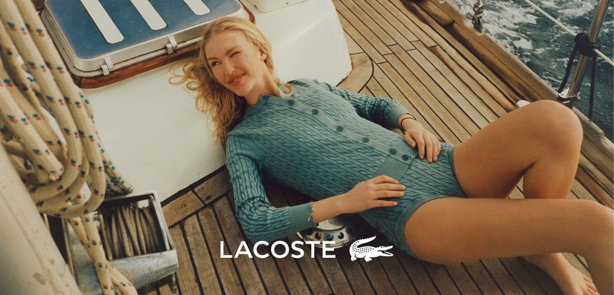 Romper lavorato a maglia azzurro chiaro con un motivo a trecce, chiusura con bottoni sul davanti e maniche corte, abbinato a un ponte in legno e corde da vela sullo sfondo.