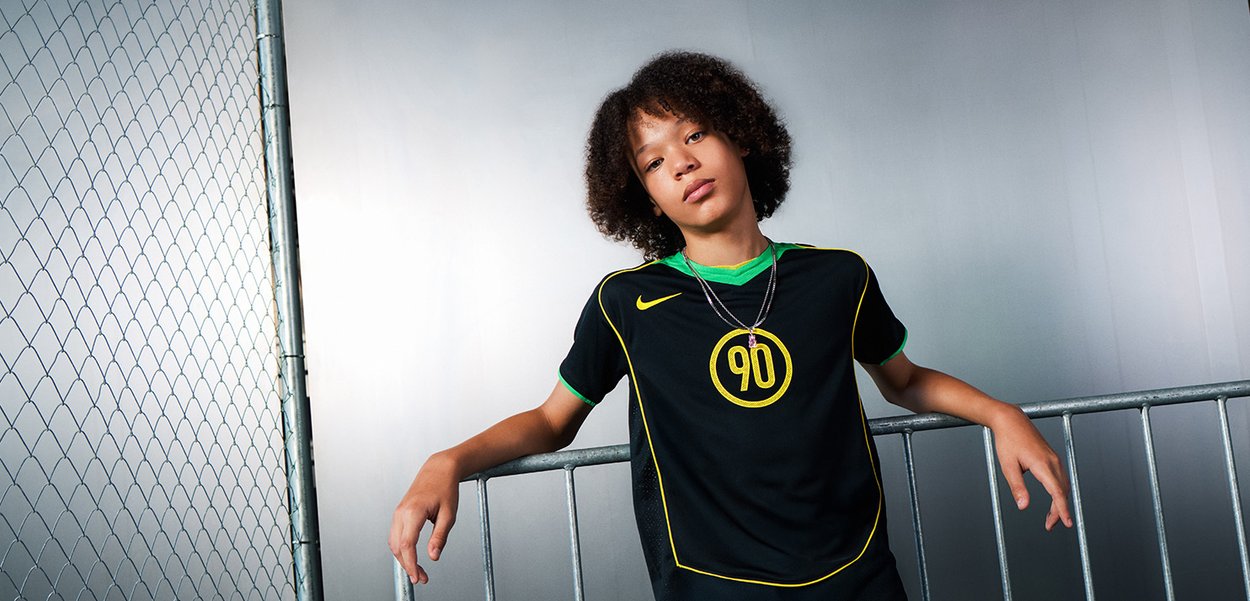 Young person with curly hair wearing a black and green sports jersey with number 90 leaning on a metal fence against a plain wall.