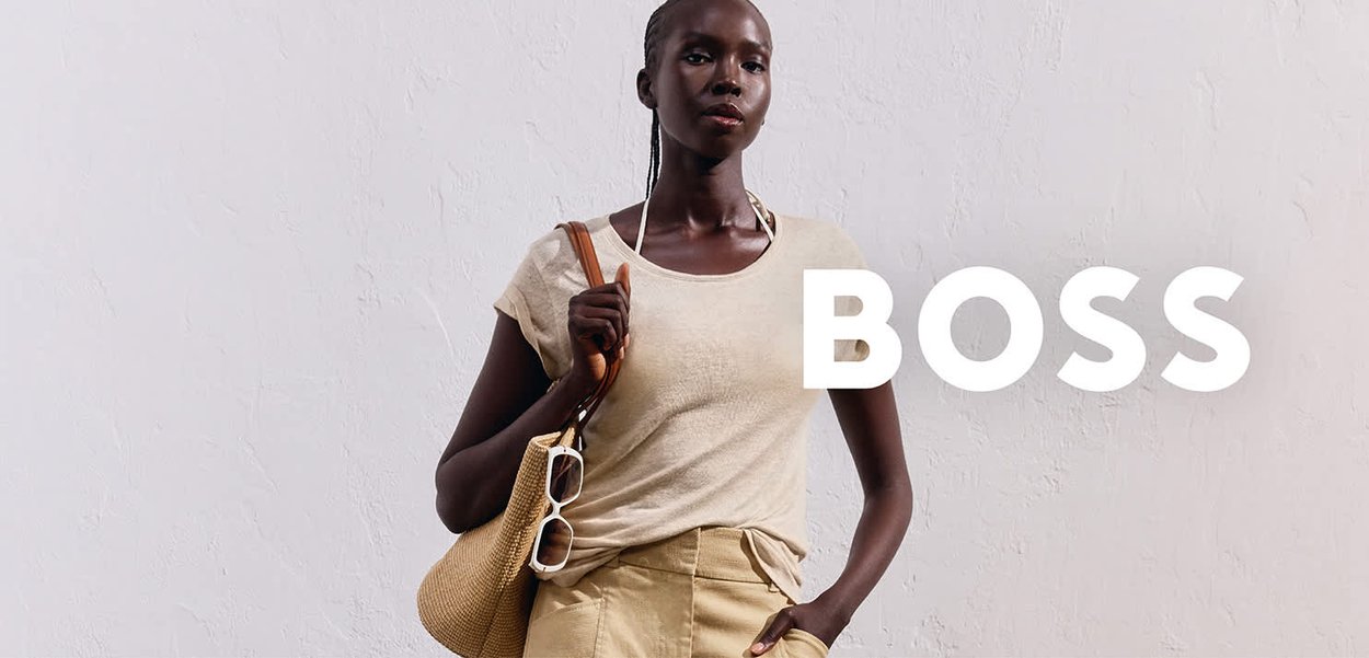 Beige Kurzarmoberteil kombiniert mit hellbraunen Hosen, hält eine gewebte Tragetasche und weiße Sonnenbrille. Einfaches, lässiges Design mit weichen Texturen.