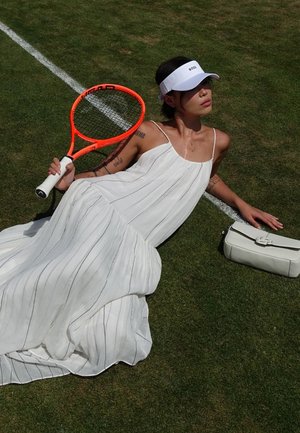 Vestito bianco senza maniche, plisettato, con sottili strisce nere; la modella tiene in mano una racchetta da tennis arancione, indossando un visiera bianca, su erba. Borsa bianca nelle vicinanze.