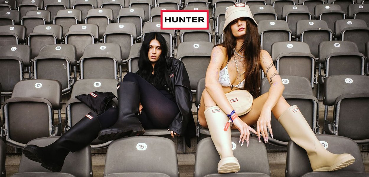 Black and beige rubber boots with logo accents, seated on stadium seats. One model wears a black jacket, the other a beige bucket hat.