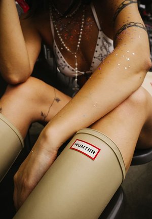 Beige rubber knee-high boots with a textured top edge and a red branded patch reading "HUNTER." Glitter accents on skin are visible.
