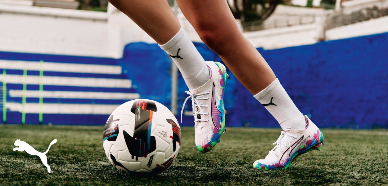 Puma soccer cleats with a predominantly white upper featuring colorful accents in pink and turquoise, paired with white socks, next to a patterned soccer ball.