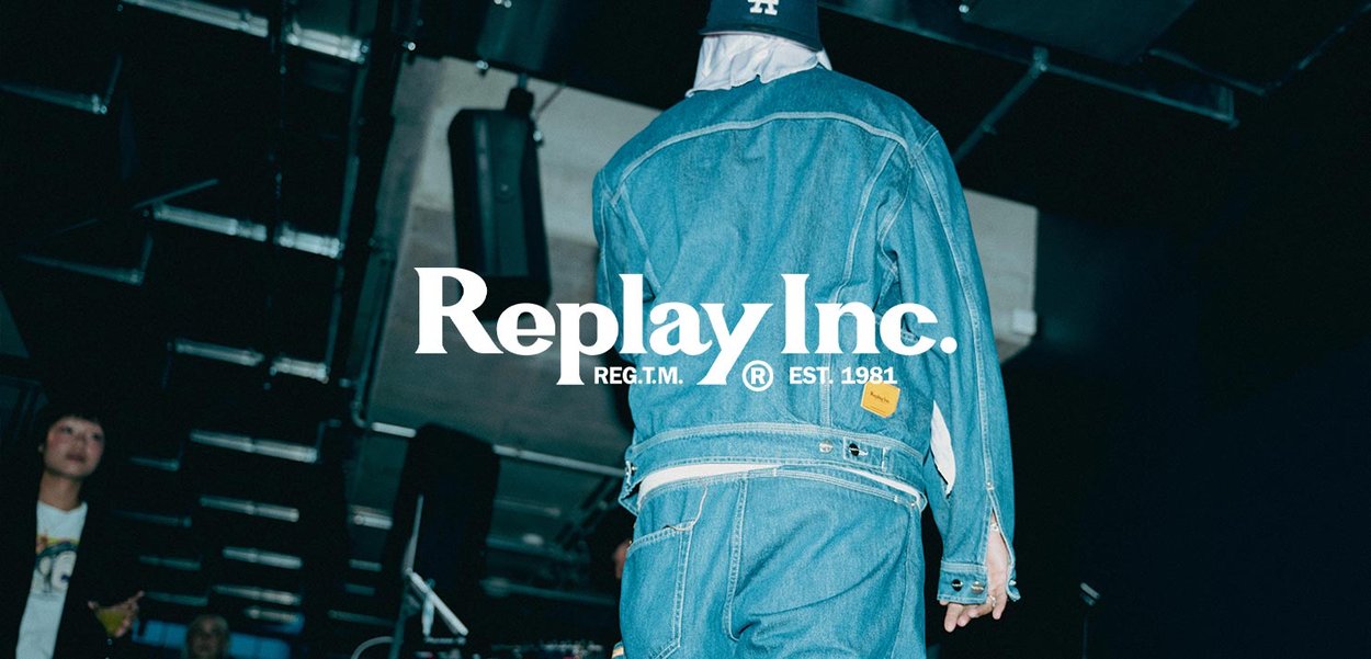 Person med en denimjacka och jeans som går inomhus under industriell takbelysning, med "Replay Inc." logotyp och textöverlägg.