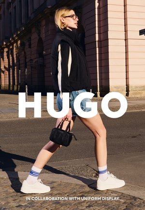 Nero gilet senza maniche con strisce bianche, pantaloni di jeans corti, sneaker bianche e borsa nera. Modello con calzini a righe blu. Sovrapposizione di testo presente.