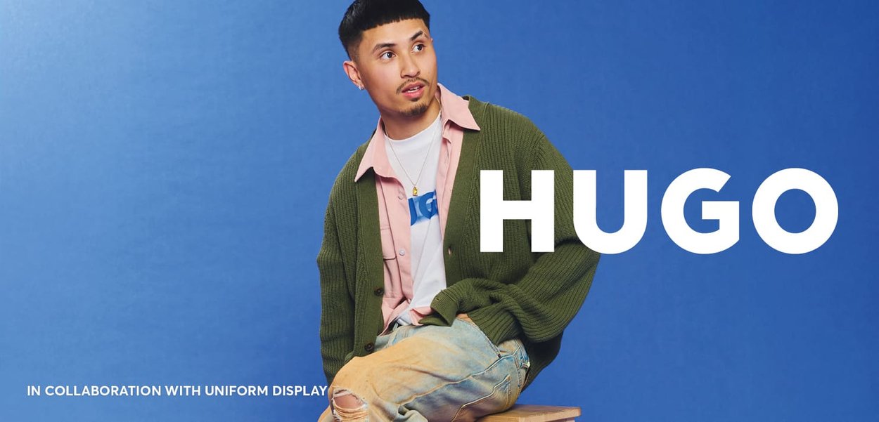 Knit green cardigan over a pink button-up shirt and white t-shirt, paired with blue denim jeans. Background is solid blue. Brand name present.