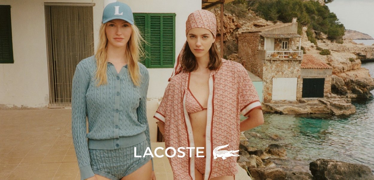 Dos mujeres posan junto a una costa rocosa. Una lleva un conjunto de suéter de punto en azul claro y una gorra, mientras que la otra viste un traje de baño con un patrón en rosa y un pareo a juego.
