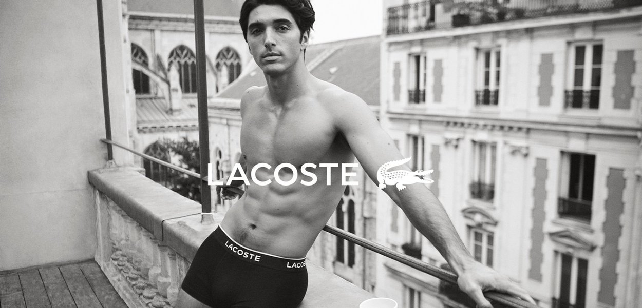 Schwarze Lacoste Boxershorts mit Logo-Bund, an einem Modell vor einem Balkon-Hintergrund präsentiert, zeigen ein minimalistisches Design und eine perfekte Passform.