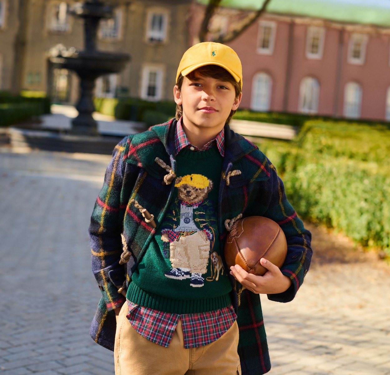 Enfant portant un manteau à carreaux avec des attaches, un pull vert avec un motif d'ours, une chemise à carreaux rouges, un pantalon beige, tenant un ballon de football marron, casquette jaune.