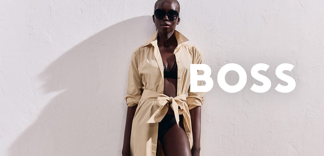 Beige button-up shirt with a tie waist, layered over a black bra. Model wearing oversized sunglasses, against a textured white background.