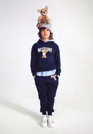 Haut à capuche bleu marine et pantalon de survêtement avec des graphiques d'ours, superposé sur une chemise rayée, complété par une casquette avec un accent d'ours et des baskets blanches.