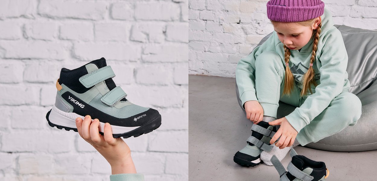 Ein Kind trägt eine lila Mütze und ein grünes Outfit und bindet sich graue und schwarze Velcro-Sneaker zu, während es auf einem silbernen Sitzsack sitzt.