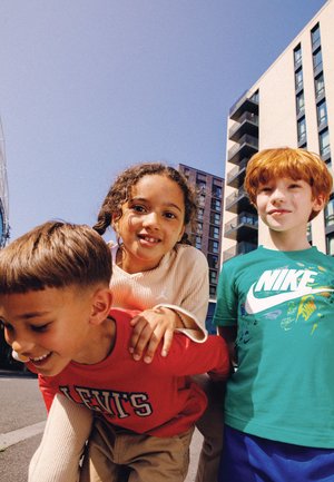 Tre bambini sorridenti all'aperto davanti a edifici residenziali moderni sotto un cielo blu chiaro in una giornata di sole.