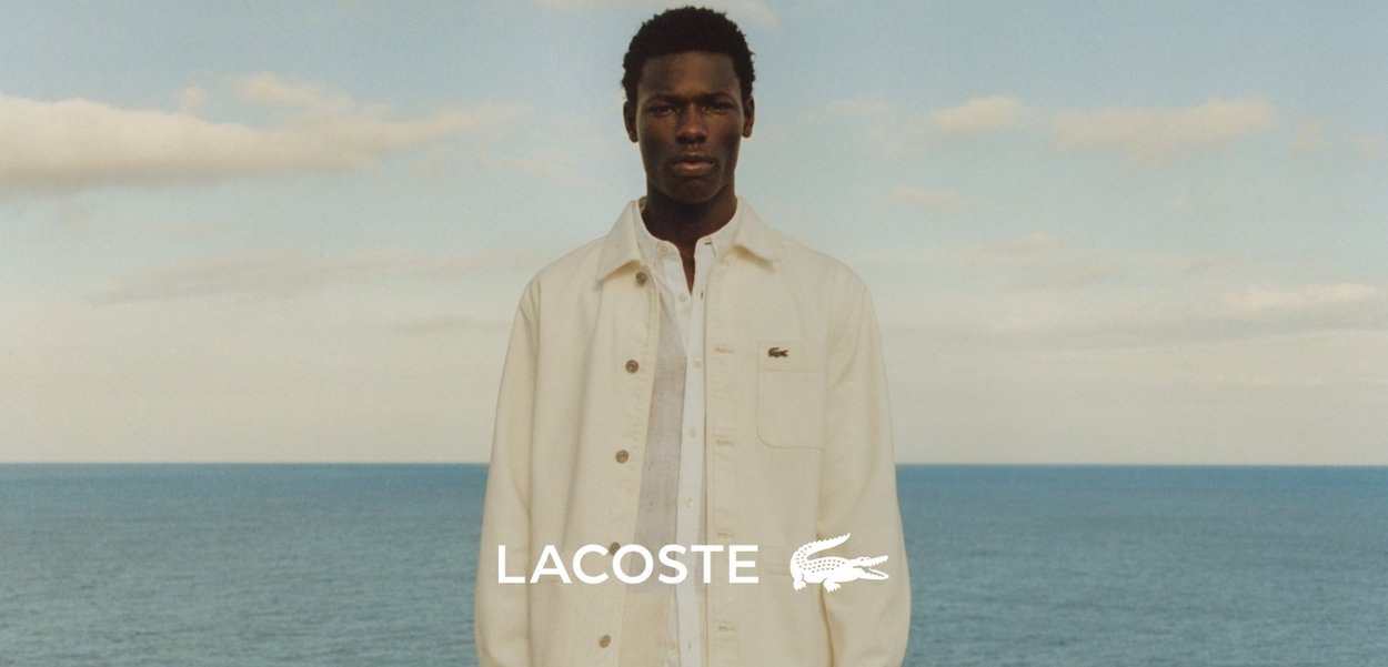 Chaqueta blanca Lacoste con cuello clásico, cierres de botón y acento de logo. El modelo está de pie frente a un océano azul bajo un cielo nublado.