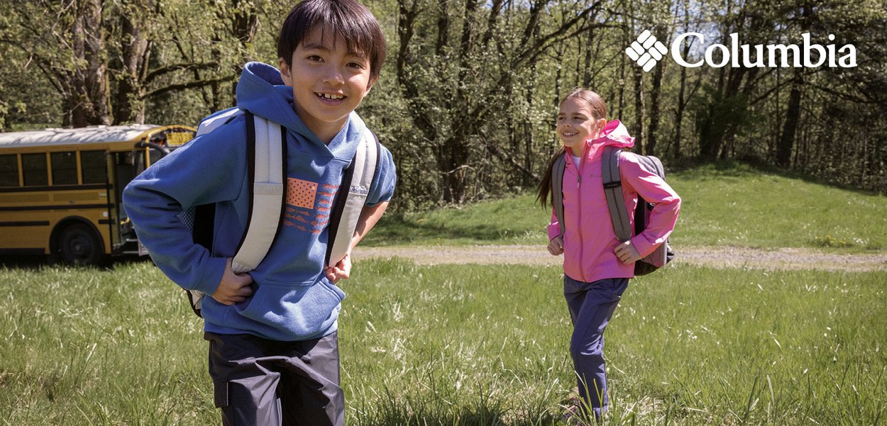 Two children run on grass, wearing a blue hoodie with an orange pattern and a pink jacket. Both have gray backpacks. A yellow school bus is in the background.