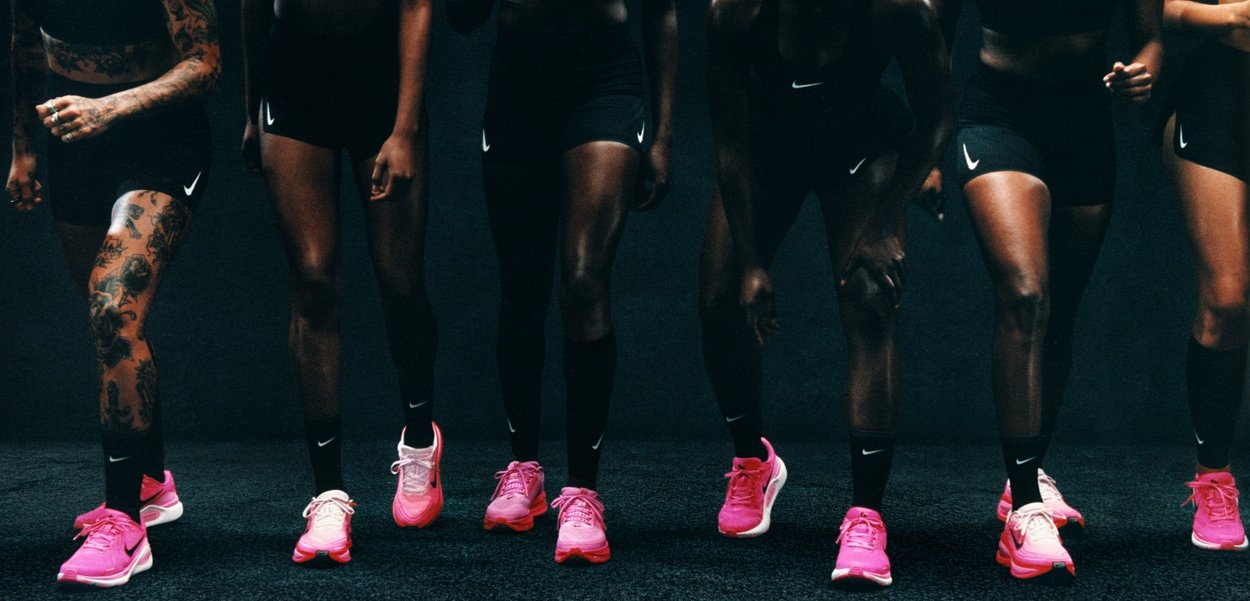 Sechs Beinpaare in schwarzen Sportshorts, die leuchtend pinke Nike-Laufschuhe mit schwarzen Akzenten tragen, vor einem dunklen Hintergrund.