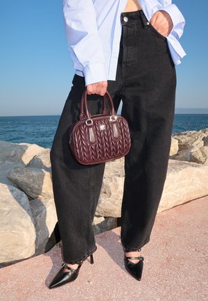 Burgundy quilted handbag with dual handles and a silver logo, paired with high-waisted black jeans and pointed black heels, by the seaside.