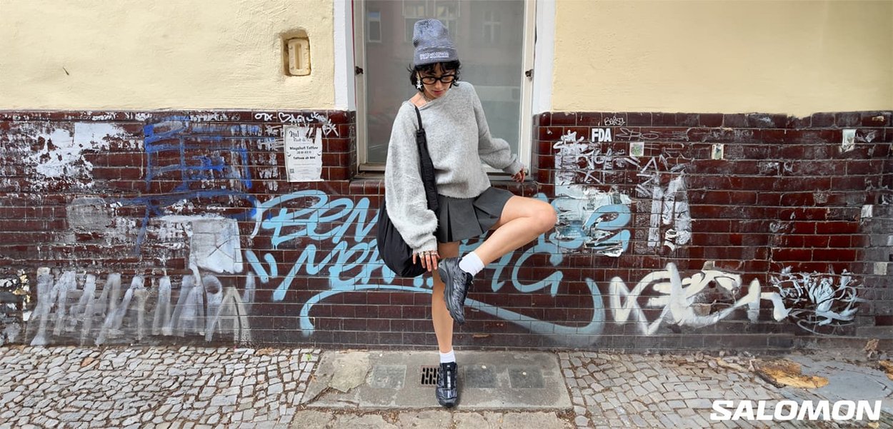 Person in gray sweater, skirt, and beanie adjusts black shoe while standing on cobblestone sidewalk in front of graffiti-covered brick wall.
