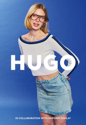 Crop top bianco a costine con dettagli blu, abbinato a una gonna in denim azzurro chiaro con un motivo a logo. Sfondo blu.