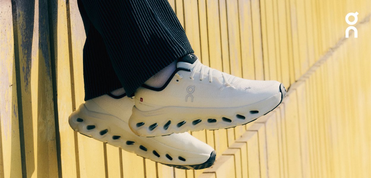 Sneakers bianchi con soletta ammortizzata, dettagli neri e logo. Parte superiore testurizzata e design traspirante. Posizionati su uno sfondo giallo.