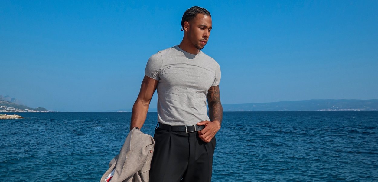 Gråtillpassad t-shirt med kort ärm, svart bälte och svarta byxor; står vid havet med en ljusgrå jacka hängande �över ena armen.