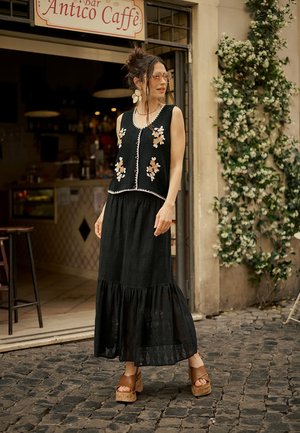 Woman in black floral vest and skirt with brown platform sandals standing outside a café with flowering vines on stone pavement.