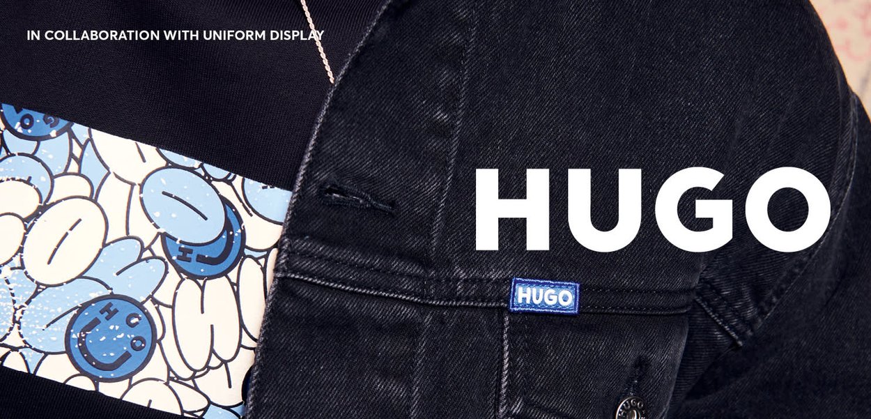 Schwarze Denimjacke mit einem blauen Logo-Patch mit der Aufschrift "HUGO" und einem bunten Grafikmuster mit lächelnden Gesichtern auf dem darunterliegenden Stoff.