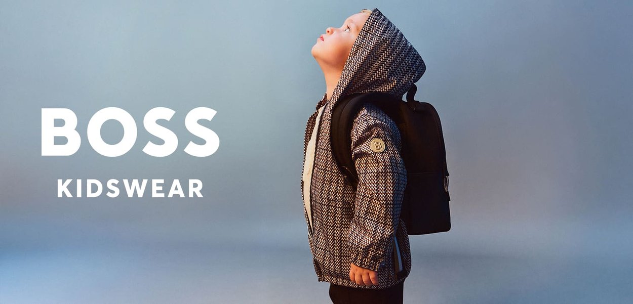 Child wearing patterned hooded jacket and black backpack looking upward against a blue background, "BOSS KIDSWEAR" text to the left.