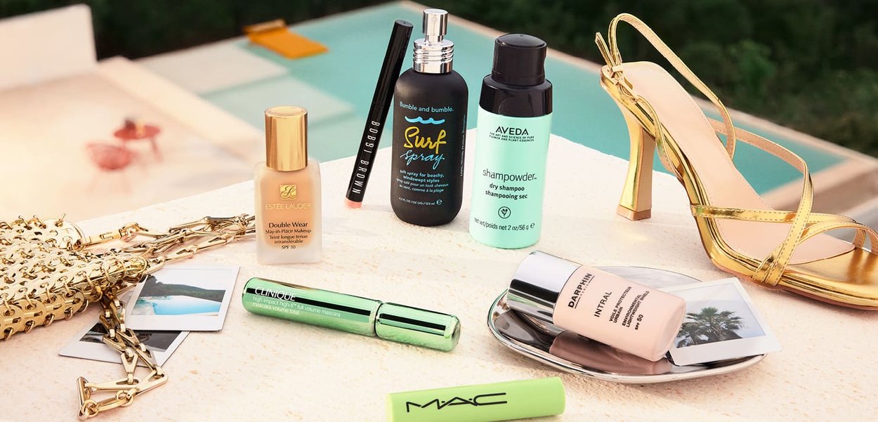 Verzameling schoonheidsproducten en een gouden hoge hak sandalen op een lichte ondergrond. De producten omvatten foundations, sprays en mascara.