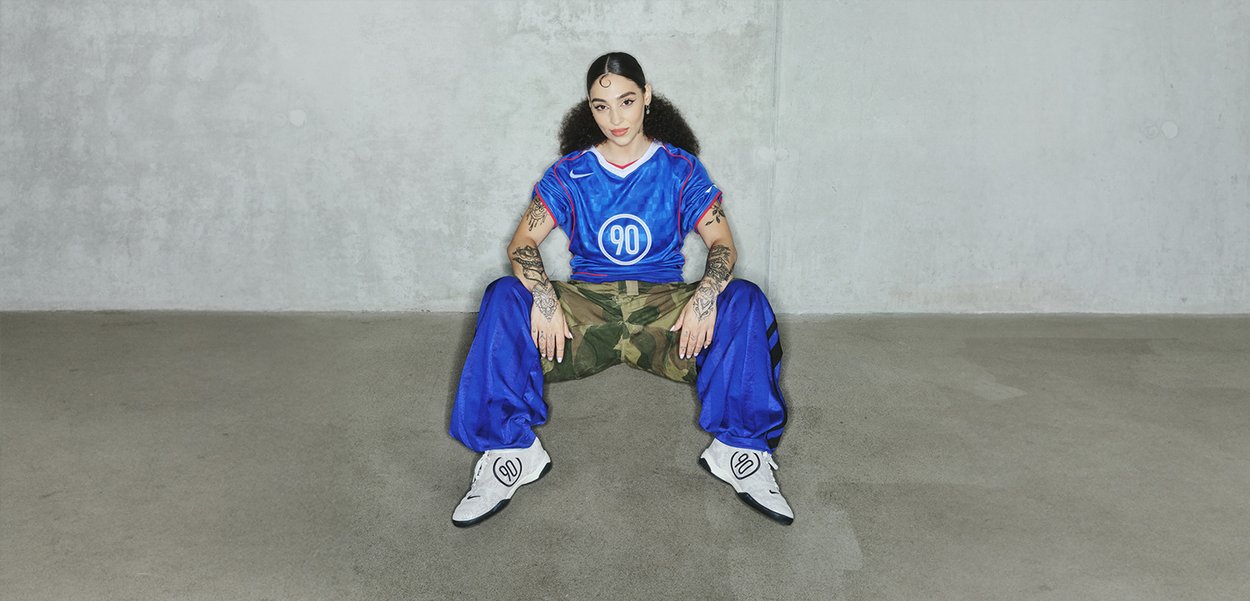 Blauw shirt met rode accenten en "90" grafiek, camouflaged pants, en witte schoenen met zwarte details, zittend tegen een grijze muur.