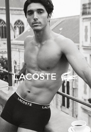 Black boxer briefs with a white "LACOSTE" logo on the waistband, worn by a person with short dark hair in a black-and-white setting.
