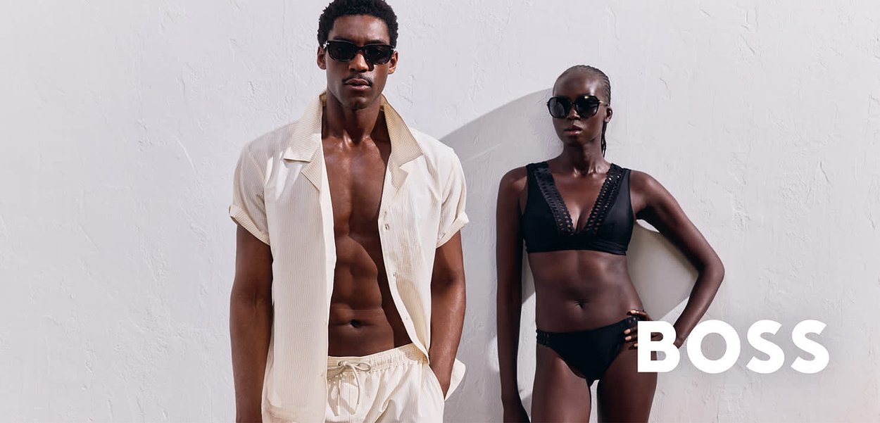 Camicia a maniche corte in crema leggera e pantaloni corti indossati da un modello maschile; la modella femminile indossa un bikini nero con rifiniture dettagliate, entrambi con occhiali da sole.