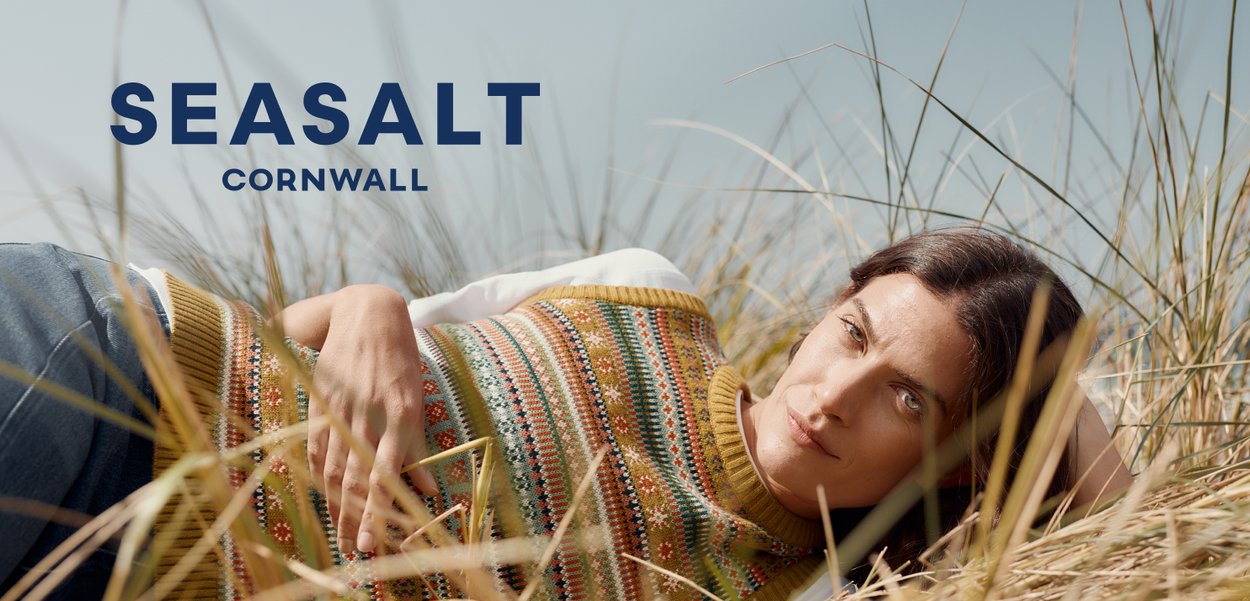Knit sweater featuring multicolored patterns in warm tones with a round neckline. Model is lying in tall grass against a light background.