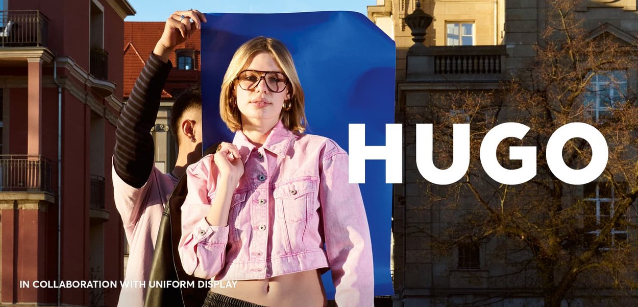Pink cropped denim jacket with metal buttons, worn over a light top. Background features a blue backdrop and urban architecture. Text: "HUGO."