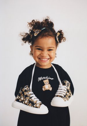 Smiling child with curly hair wearing a black Moschino shirt and holding matching teddy bear-patterned sneakers around their neck.